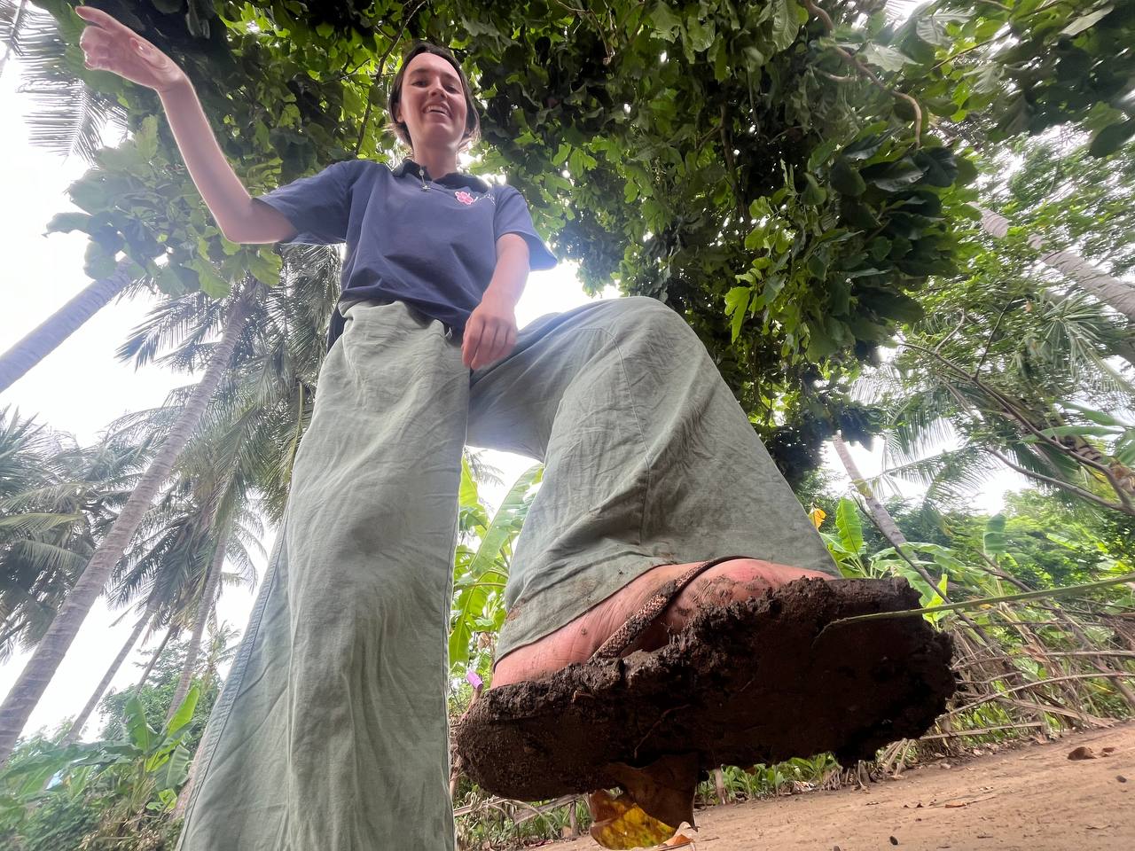 Brieya laughing and reaching into the jungle canopy while standing on a muddy path in Cambodia.