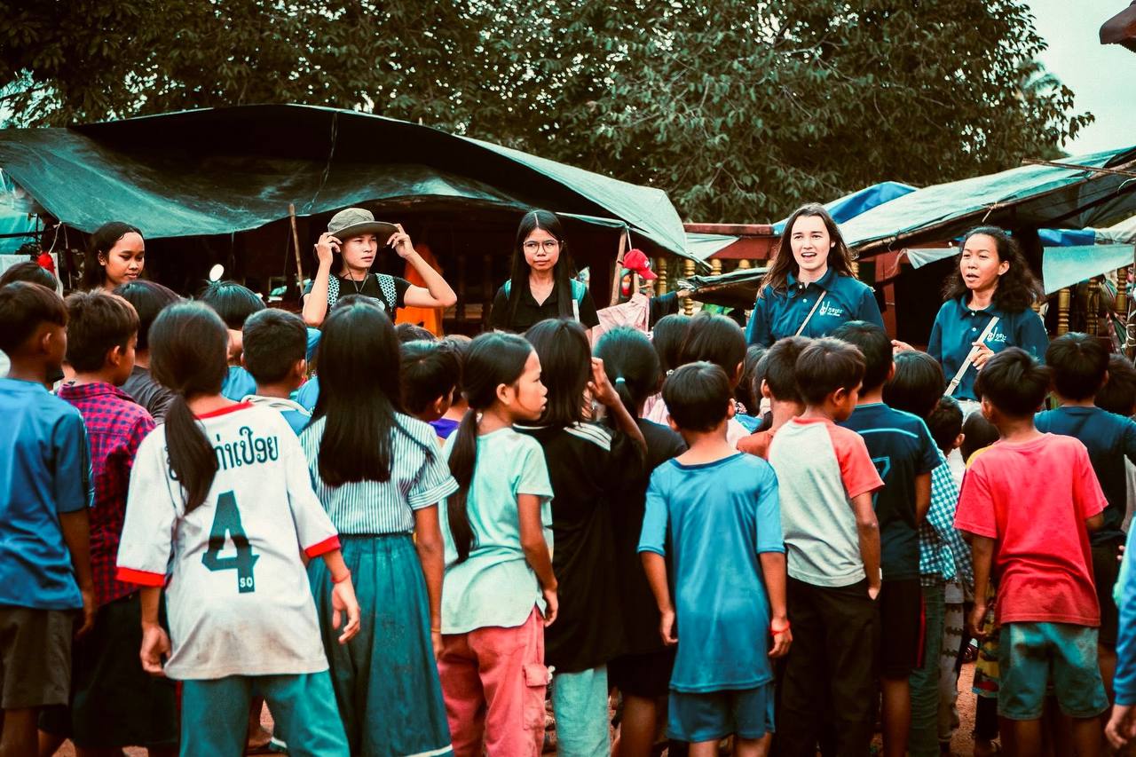 Brieya and ministry staff surrounded by a crowd of Cambodian children in a village market setting.