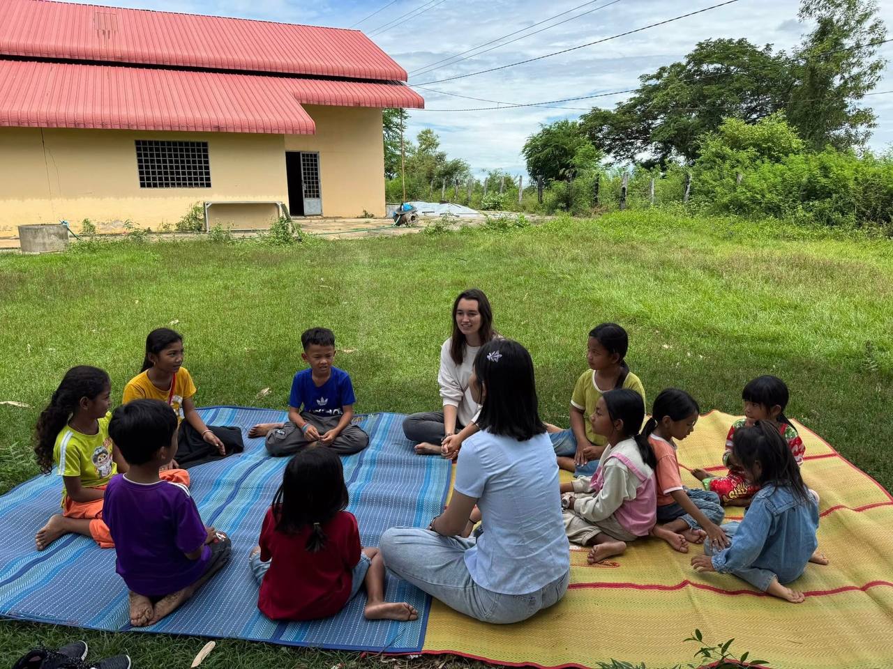 Brieya and a local staff member teaching a circle of children seated on mats outside a school building.