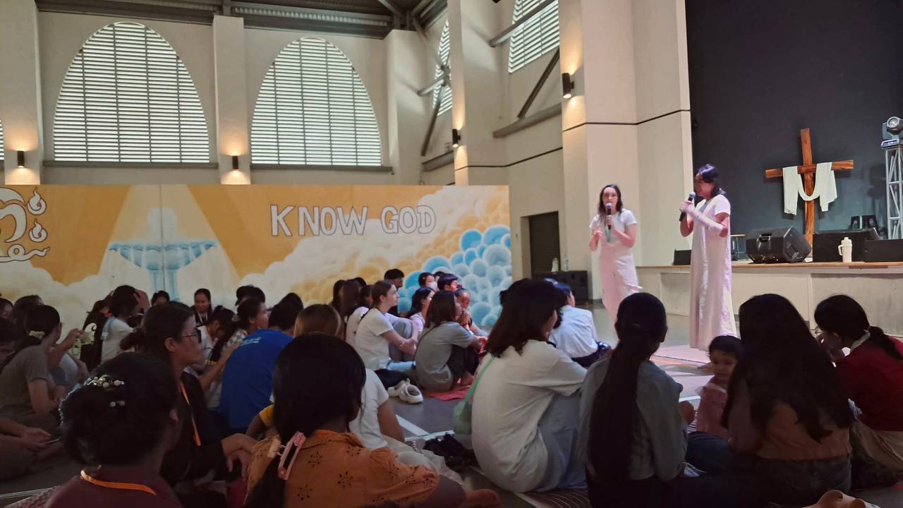 Brieya speaking on stage with a microphone in front of a 'Know God' backdrop and wooden cross.