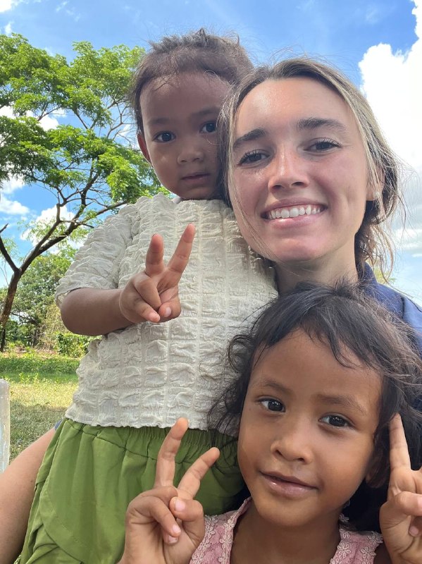 Brieya smiling closely with two young Cambodian children perched on her shoulders under a blue sky.