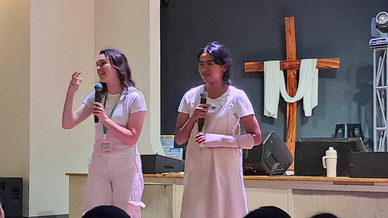Brieya on stage with a young girl, both holding microphones, in front of a large wooden cross.